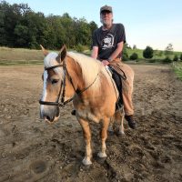 A man with a grey beard and glasses wearing a baseball cap sits atop a light chestnut horse with a white mane in a dirt riding arena.