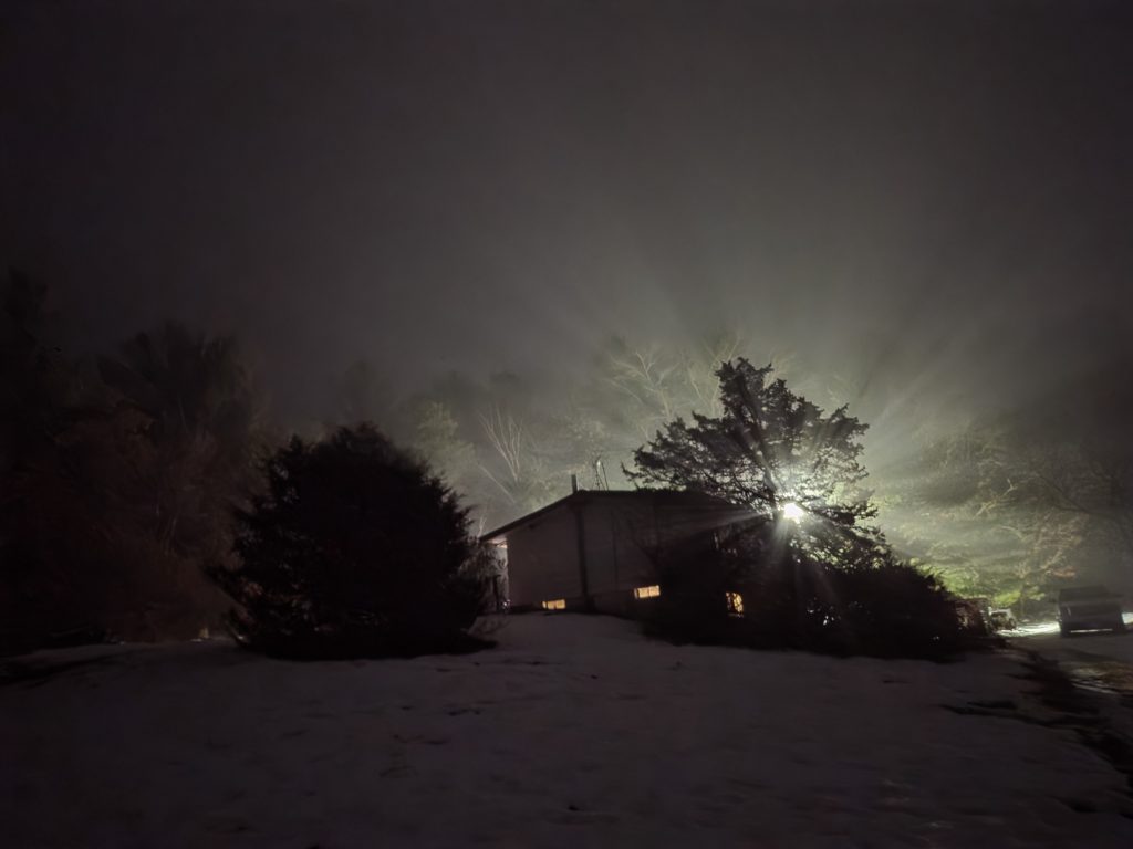 A dark, snowy backyard at night covered in thick fog, with a bright light beaming through tree branches creating dramatic rays of light.