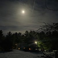 Full moon over a snowy yard in Otter Creek at night, showing deer trails in the snow, glowing house windows, and a vapor trail in the sky.
