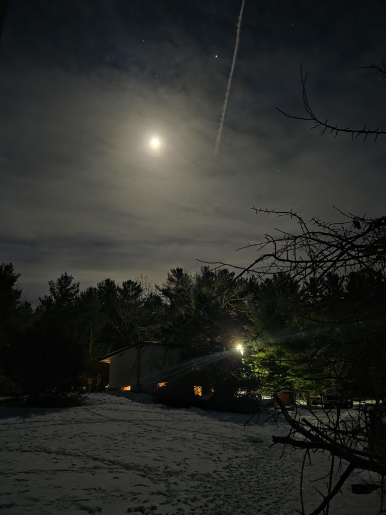Full moon over a snowy yard in Otter Creek at night, showing deer trails in the snow, glowing house windows, and a vapor trail in the sky.